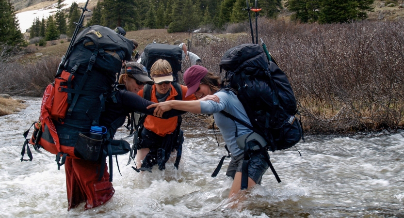 Three people wearing backpacks stand in knee-deep rushing water, bracing themselves with each others' arms. 